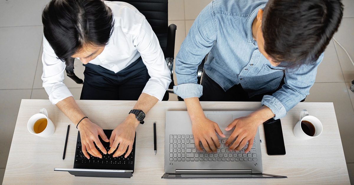 two people typing on separate laptops at one shared desk from above