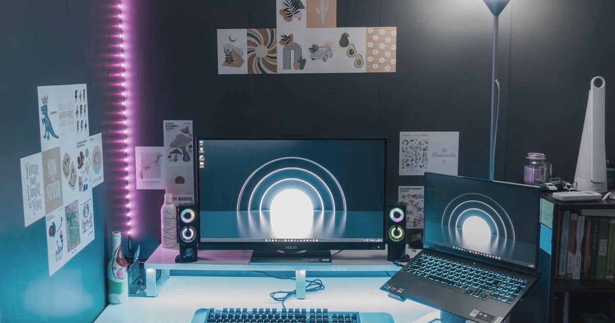 A desk with a monitor and a laptop side by side under coloured light