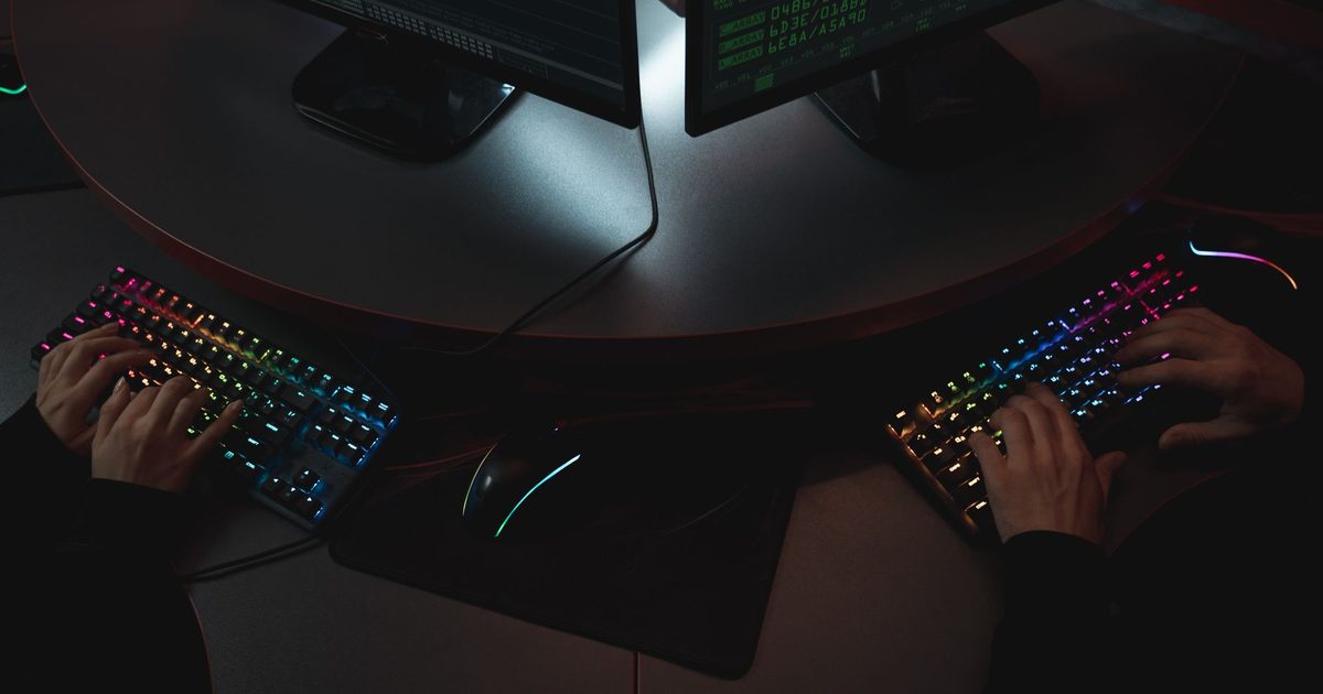 two backlit gaming keyboards and a mouse on a dark desk with monitors