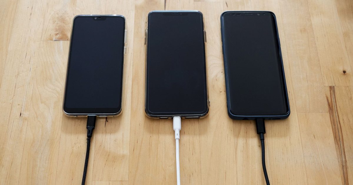 three smartphones lying on a wooden desk each plugged into a charging cable